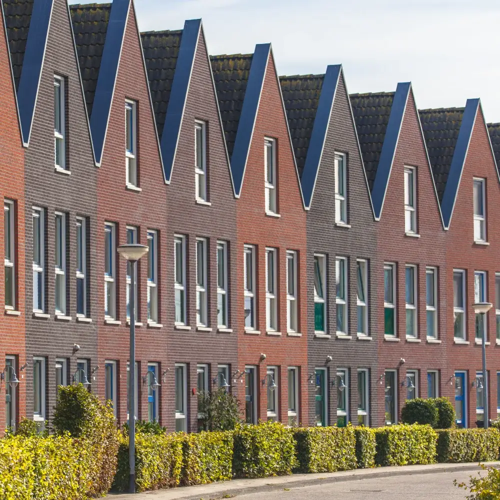 Modern brick townhouses with repeating sharp gable roofs, tall windows and green hedgerows along a residential street.